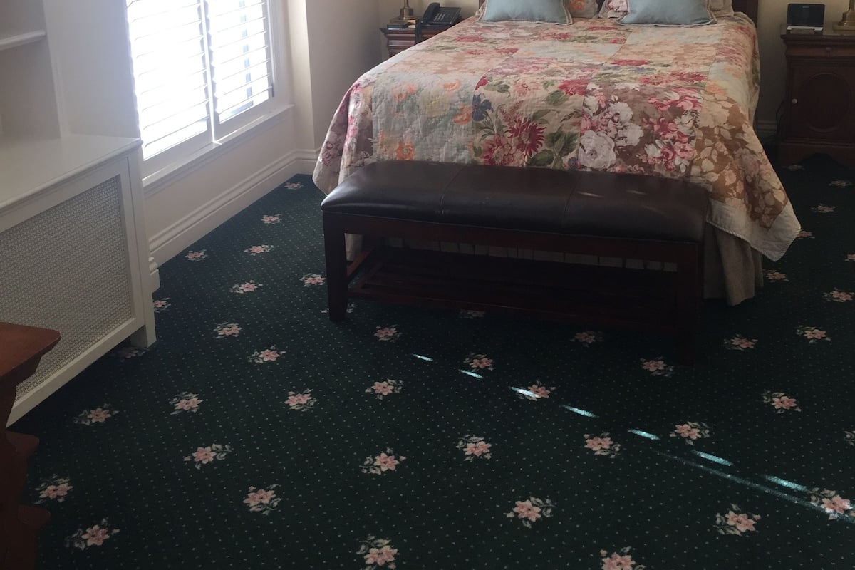 A bedroom with a floral bedspread, a wooden bench, a nightstand with a lamp, and a window with blinds.