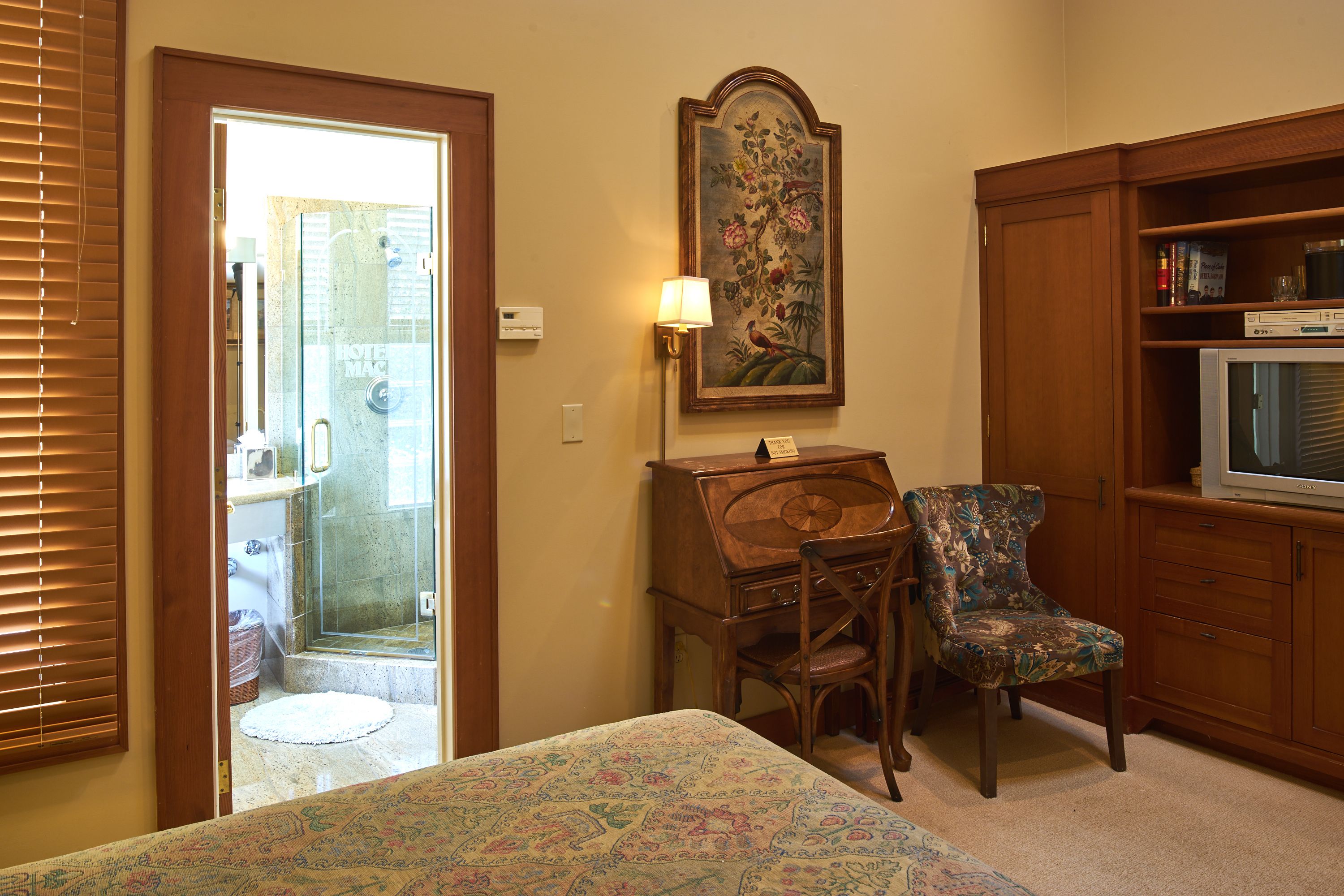 A hotel room with a bed, a wooden desk, a chair, a television, and a painting on the wall.