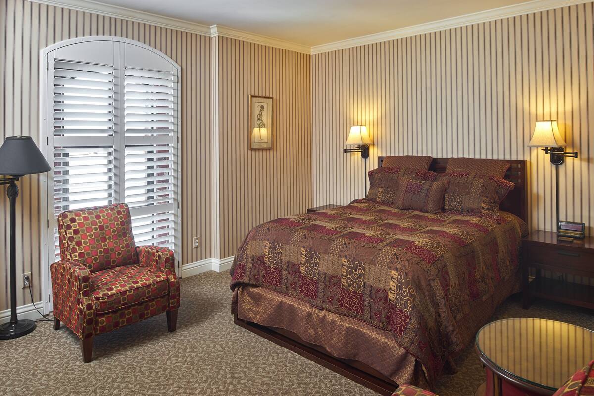 A hotel room with a large bed, a chair, a floor lamp, and a bedside table.