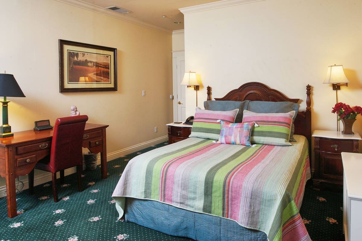 A hotel room with a bed, a desk, a chair, a lamp, and a framed picture on the wall.