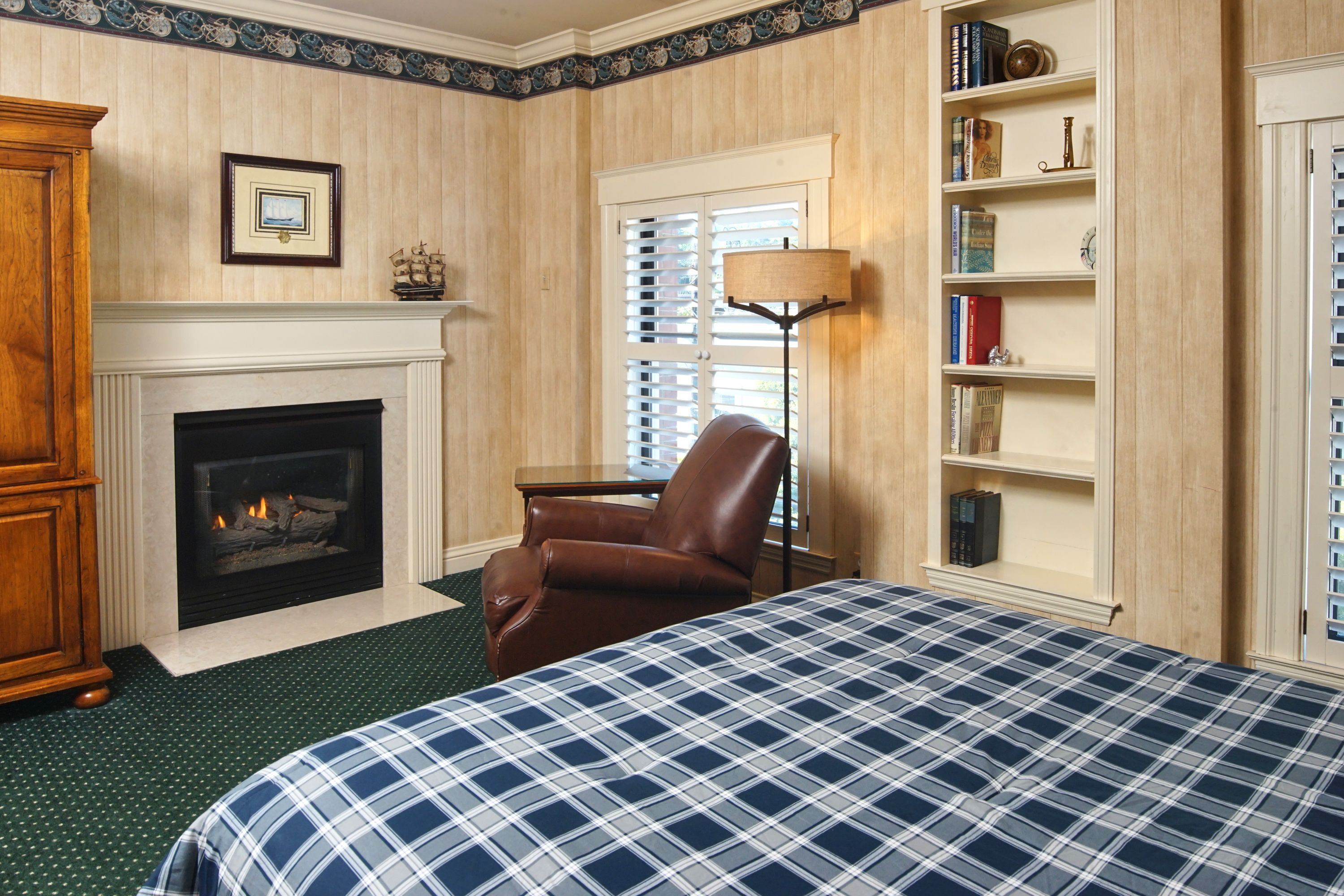 Una habitación de hotel con chimenea, una cama con colcha escocesa, un sillón de cuero, una estantería y una ventana con persianas.