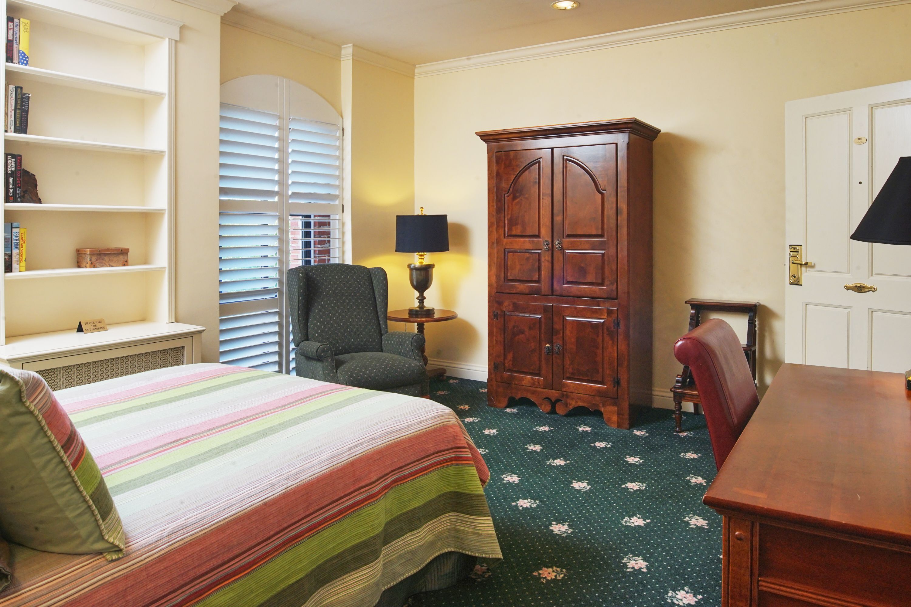 A hotel room with a bed, a chair, a wooden cabinet, a small table with a lamp, and a desk.
