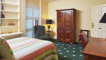 A hotel room with a bed, a chair, a wooden cabinet, a small table with a lamp, and a desk.