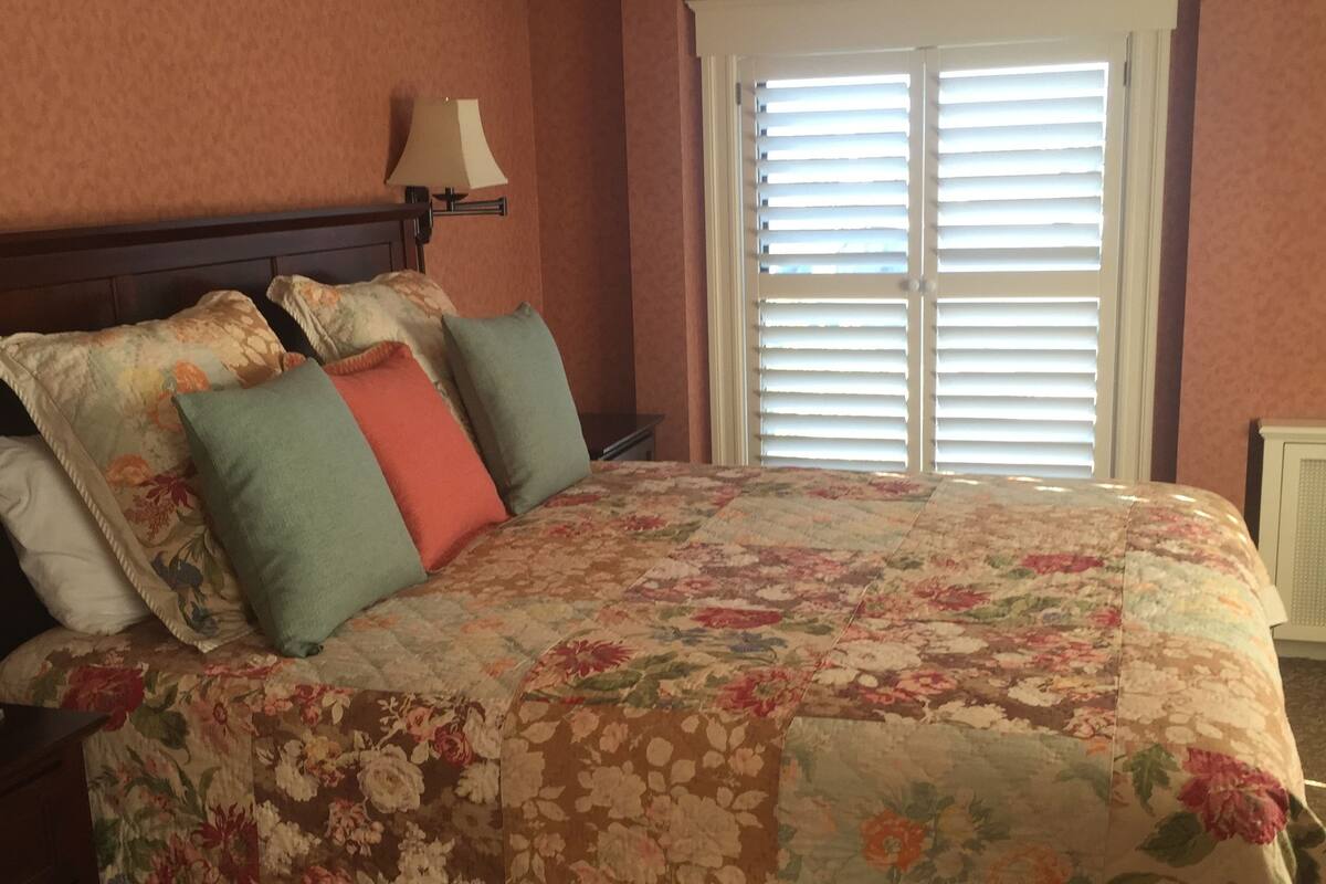 A bedroom with a floral bedspread, a nightstand with a lamp, and a window with blinds.