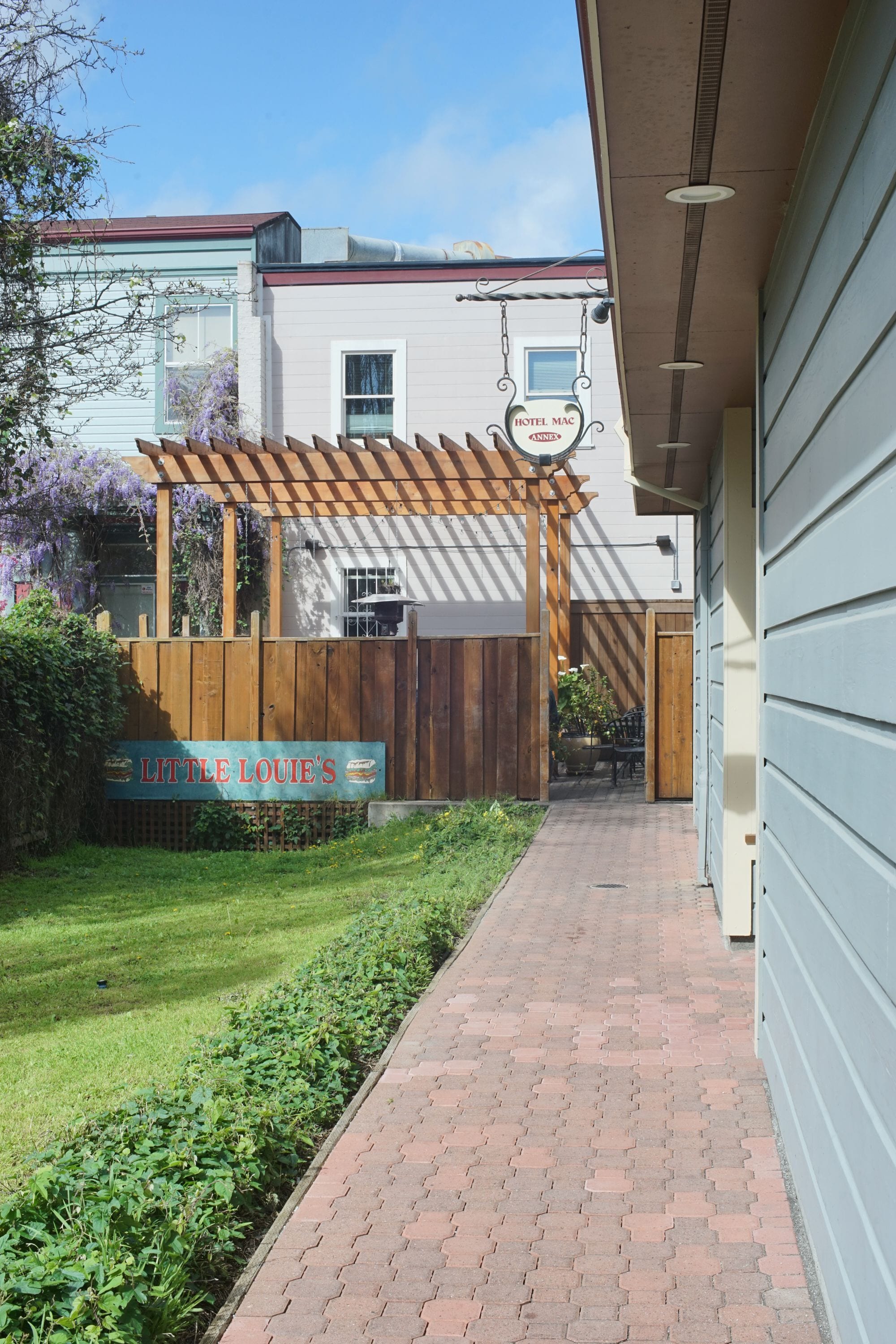 Un camino de ladrillos que conduce a una pérgola de madera con un letrero que dice 'Little Louie's' y un cartel de 'Hotel Mac' arriba.