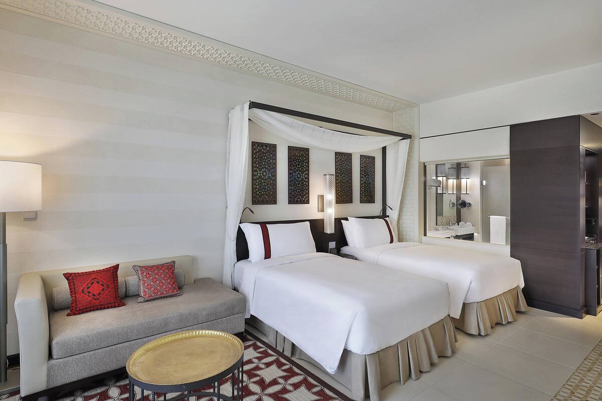 A hotel room with two beds, a sofa, a small table, and a bathroom visible through a glass partition.