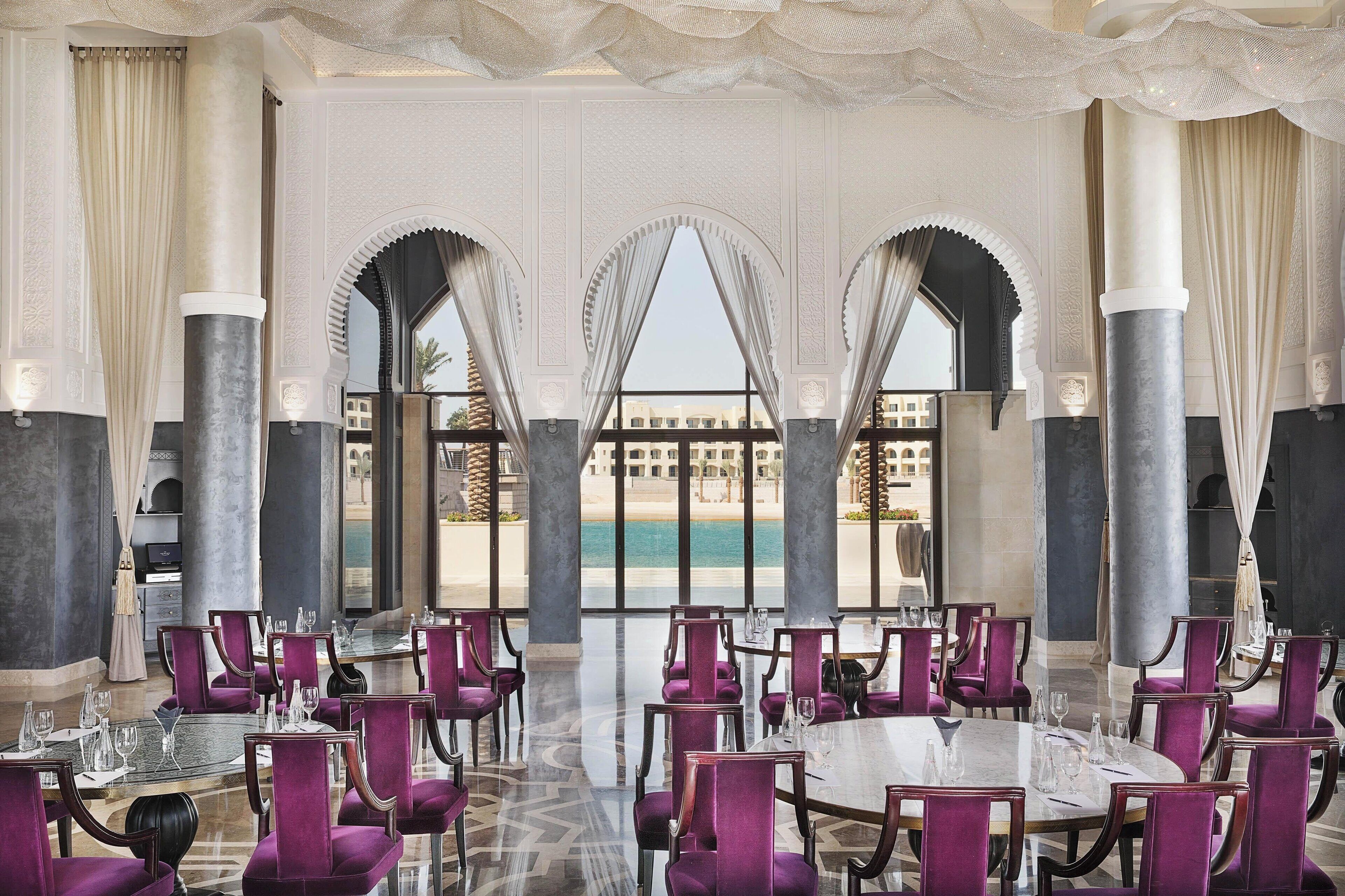 A luxurious dining area with purple upholstered chairs and tables, large columns, and a view of the beach.