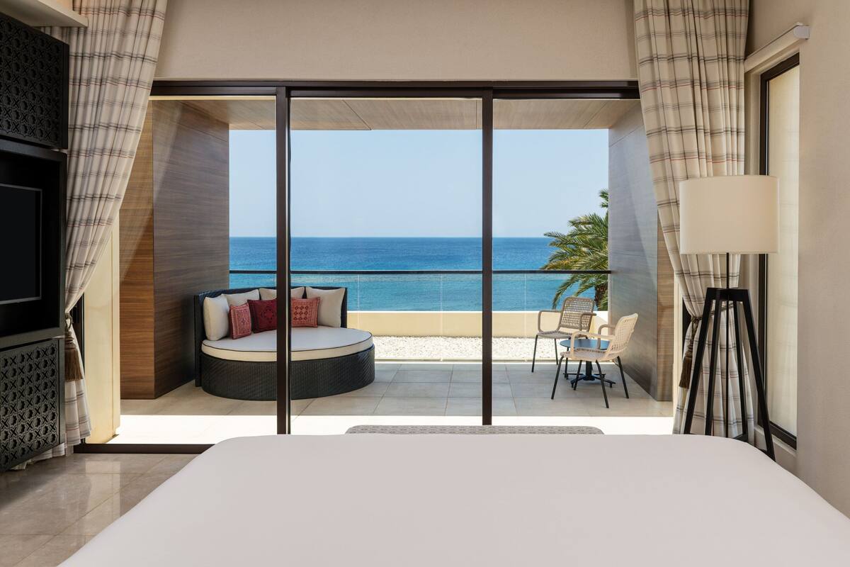 A bedroom with a large bed, a seating area with a view of the sea, and a balcony with a table and chairs.