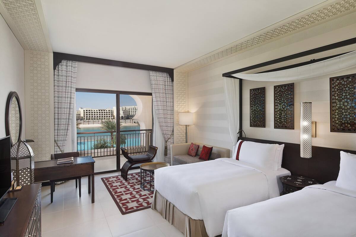 A hotel room with two beds, a desk, a chair, a TV, and a balcony with a pool view.