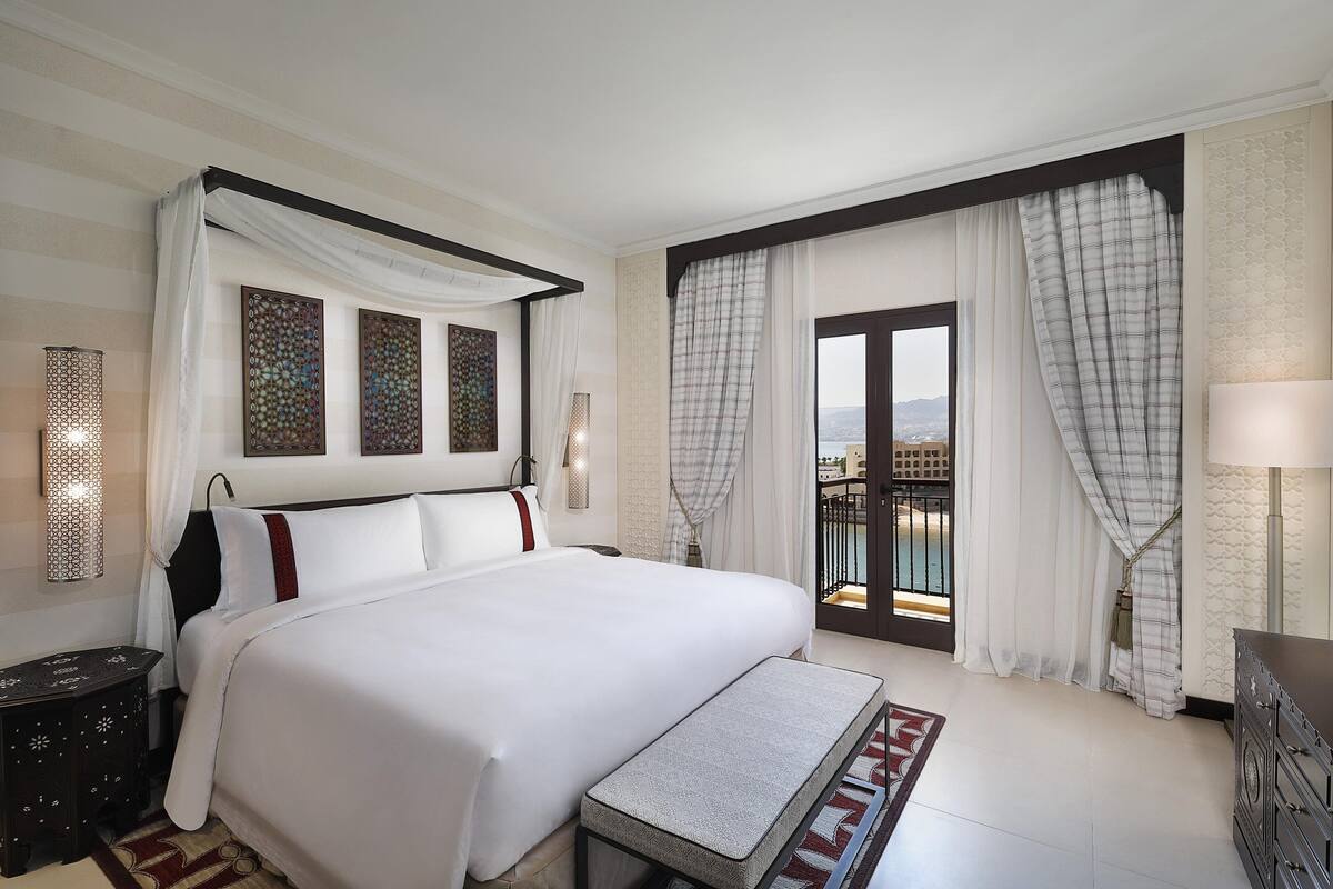 A large bed with a canopy, a bedside table, a bench, a dresser, and a view of the outdoors through a balcony.