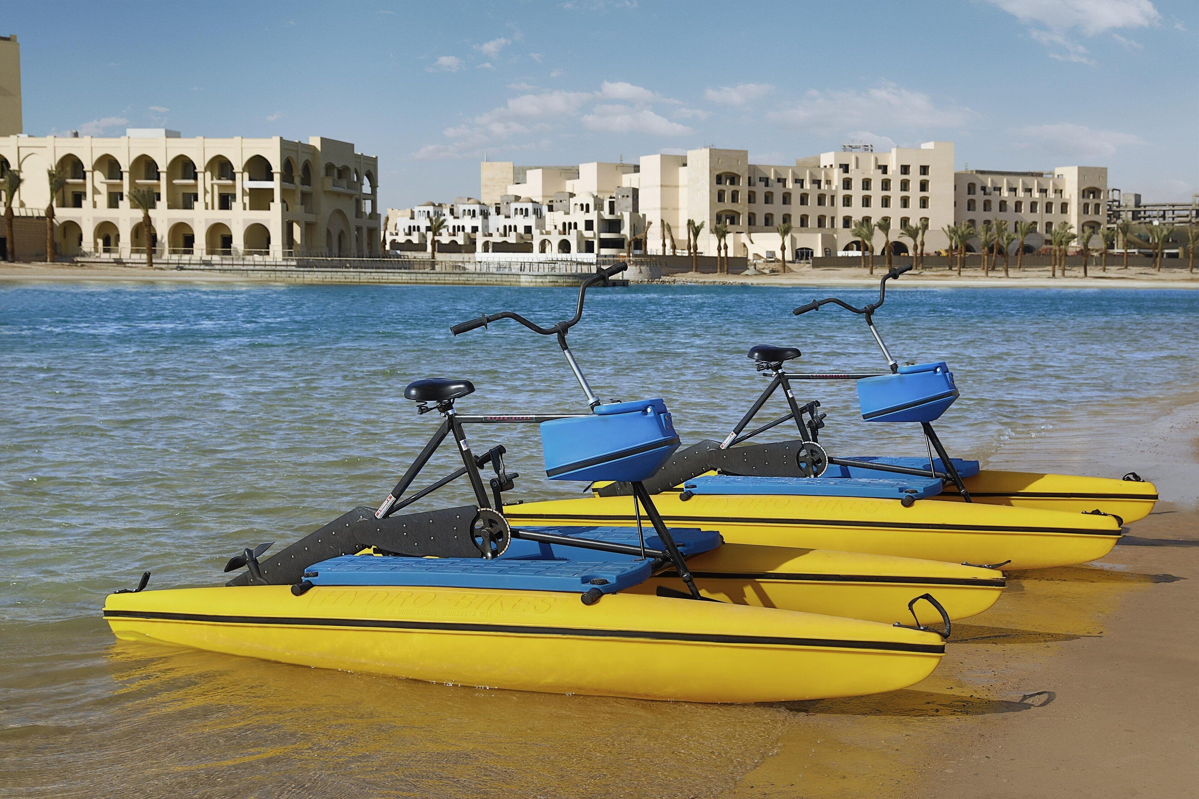 A row of yellow paddle boats with blue seats and handlebars, docked at a sandy beach with a backdrop of white buildings and palm trees.