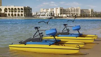 A row of yellow paddle boats with blue seats and handlebars, docked at a sandy beach with a backdrop of white buildings and palm trees.