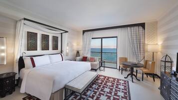 A hotel room with a large bed, a small table, a sofa, and a balcony with a view of the sea.