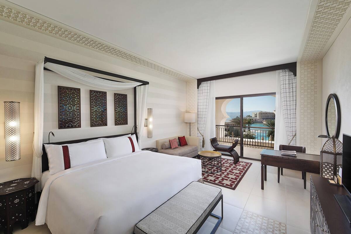 A hotel room with a large bed, a sofa, a dining table, and a balcony with a pool view.