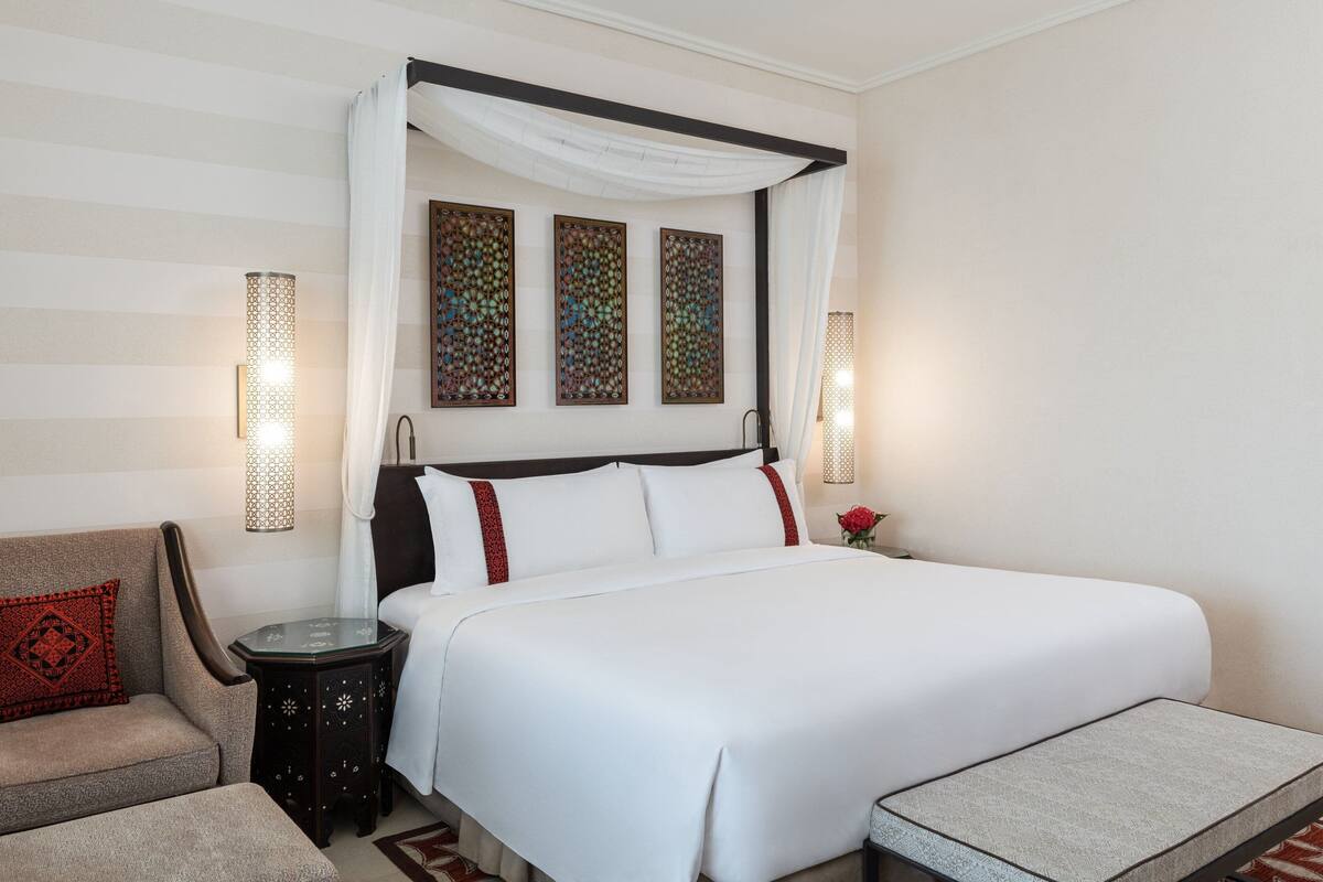 A neatly arranged hotel room with a four-poster bed, a side table, a chair, and decorative wall art.