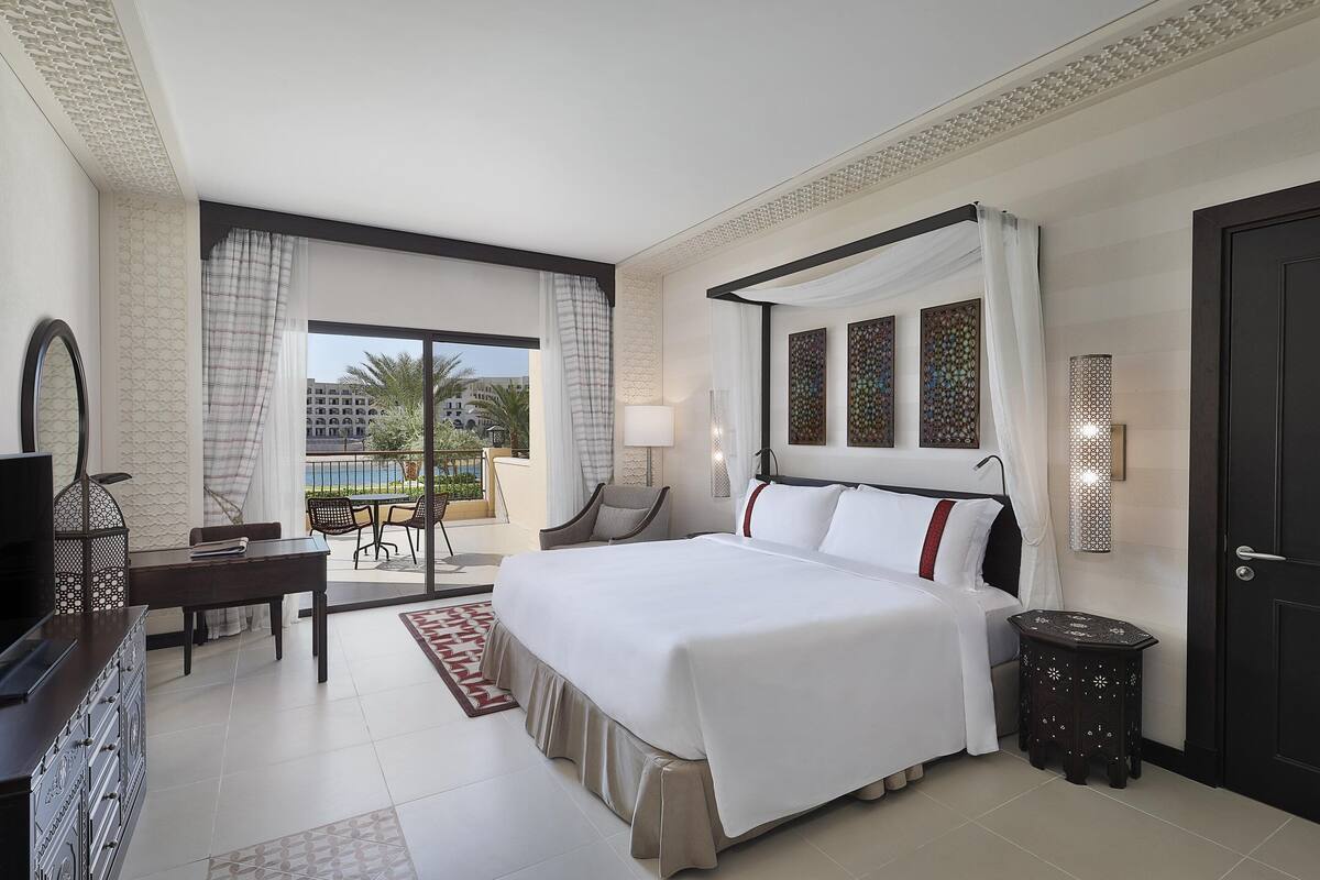 A hotel room with a large bed, a TV, a desk, and a balcony with a view of palm trees.