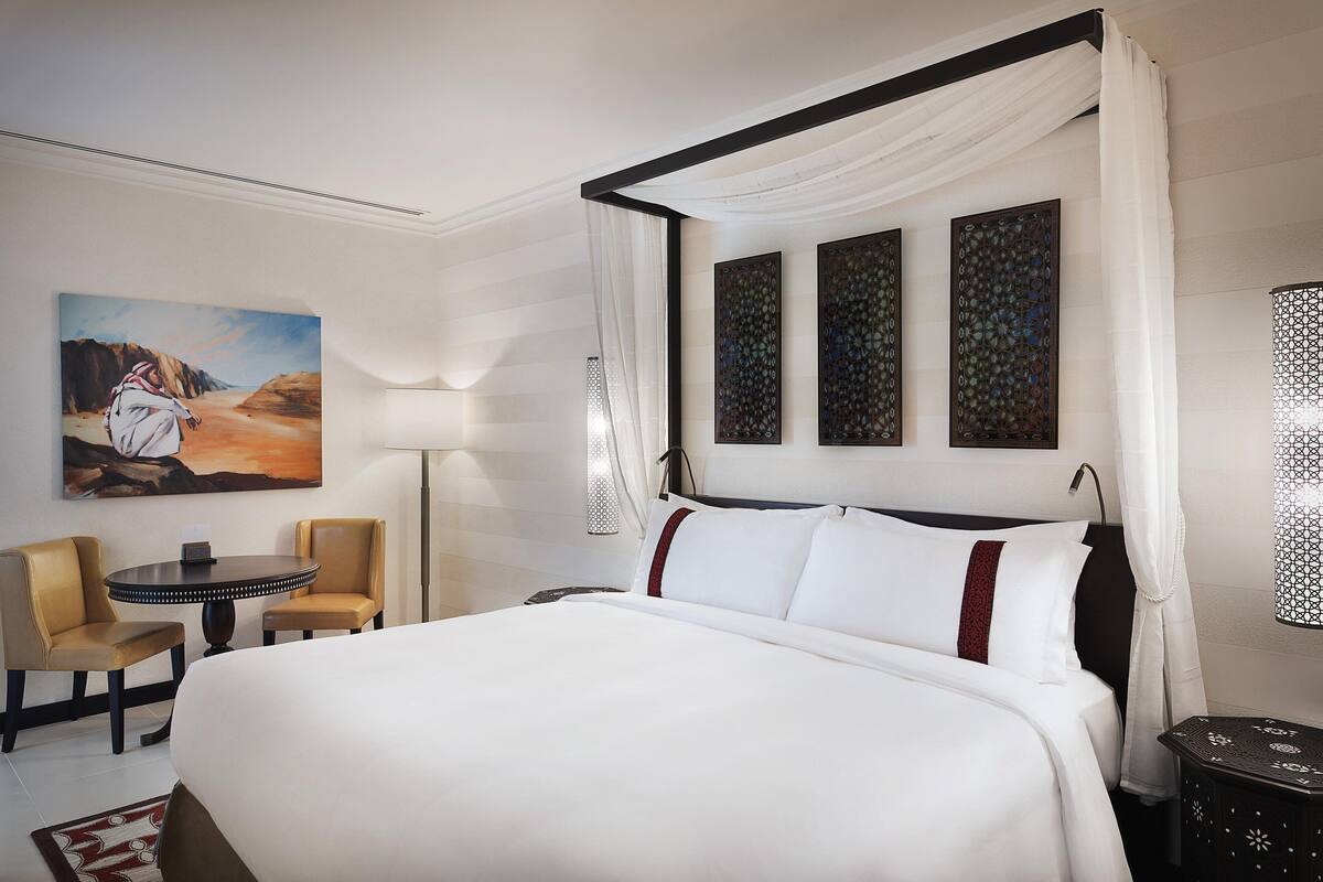 A hotel room with a large bed, two chairs, a round table, a painting, and decorative wall panels.