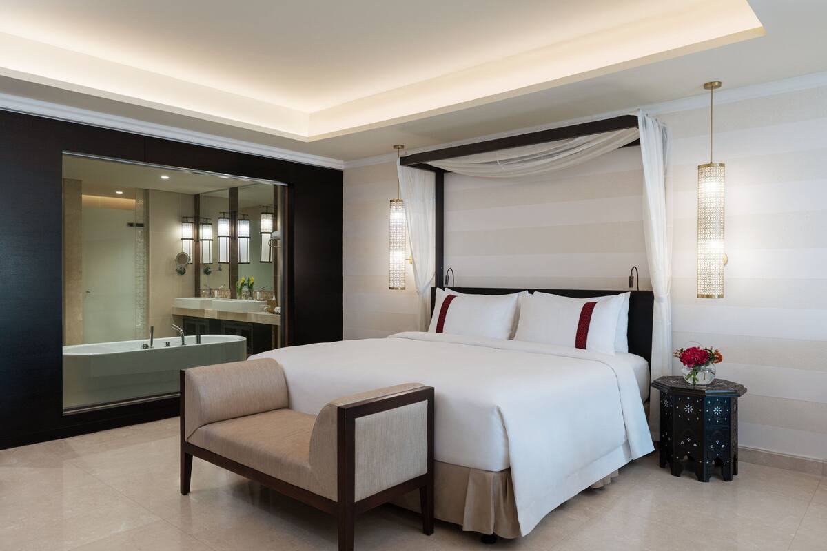 A modern hotel room with a large bed, a sitting area, a vanity with a mirror, and a large window.