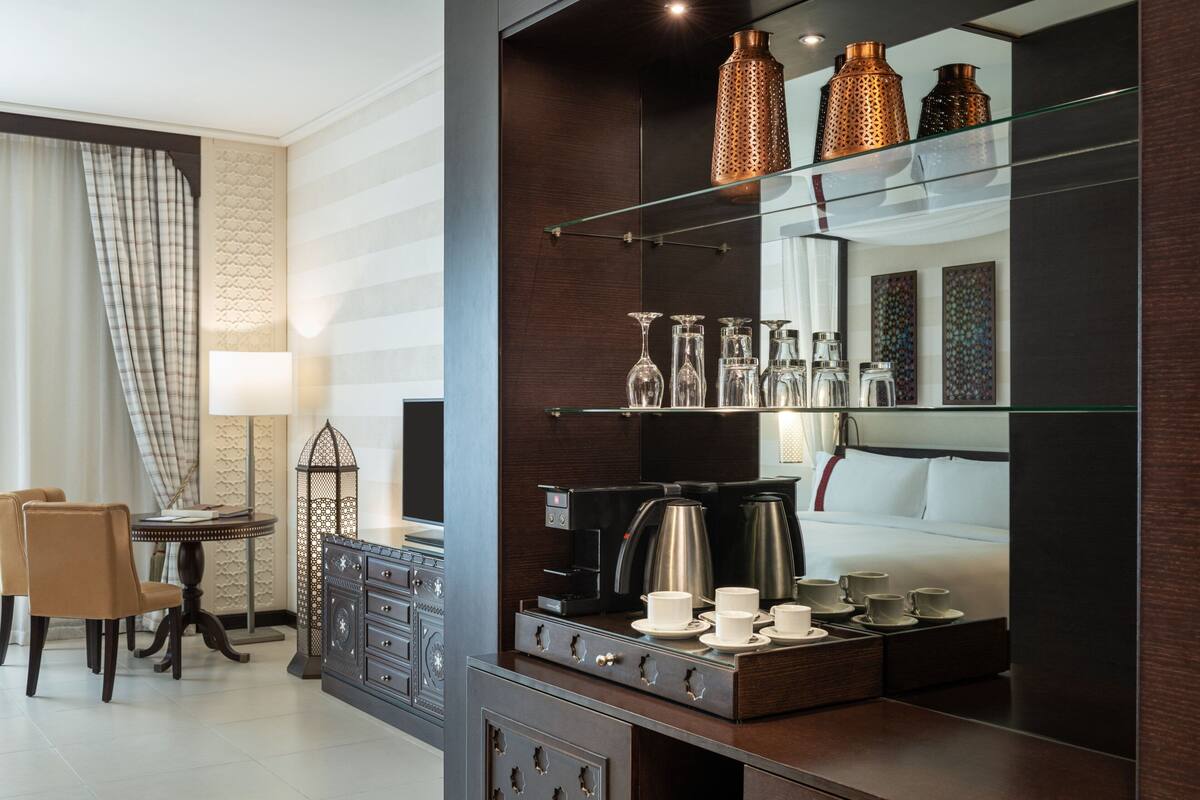 A modern hotel room with a dark wood cabinet, a glass shelf with decorative vases, a coffee station, and a dining area with a table and chairs.