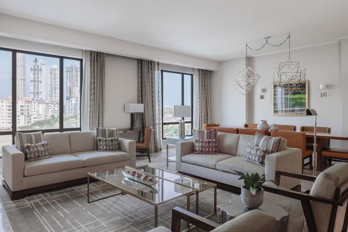 A modern living room with two sofas, a coffee table, and a dining area. Large windows offer a view of the cityscape.
