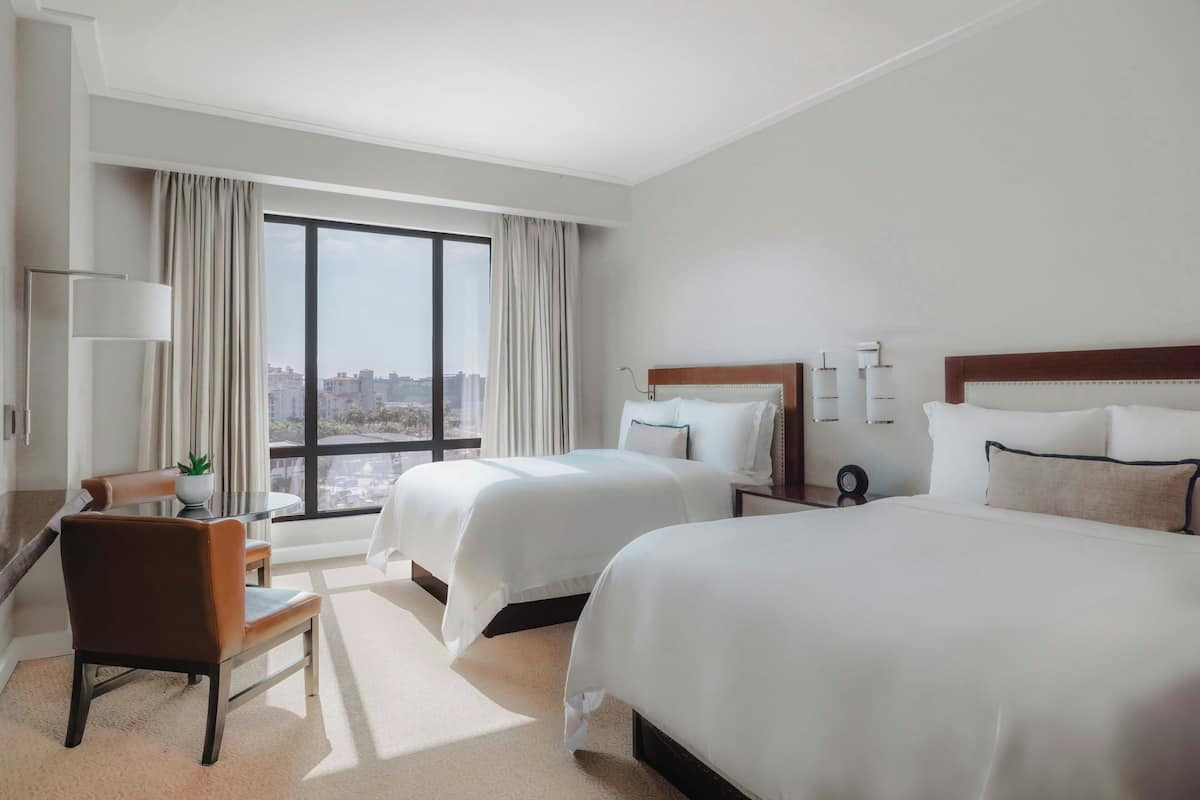 A hotel room with two beds, a desk, a chair, and a large window offering a city view.