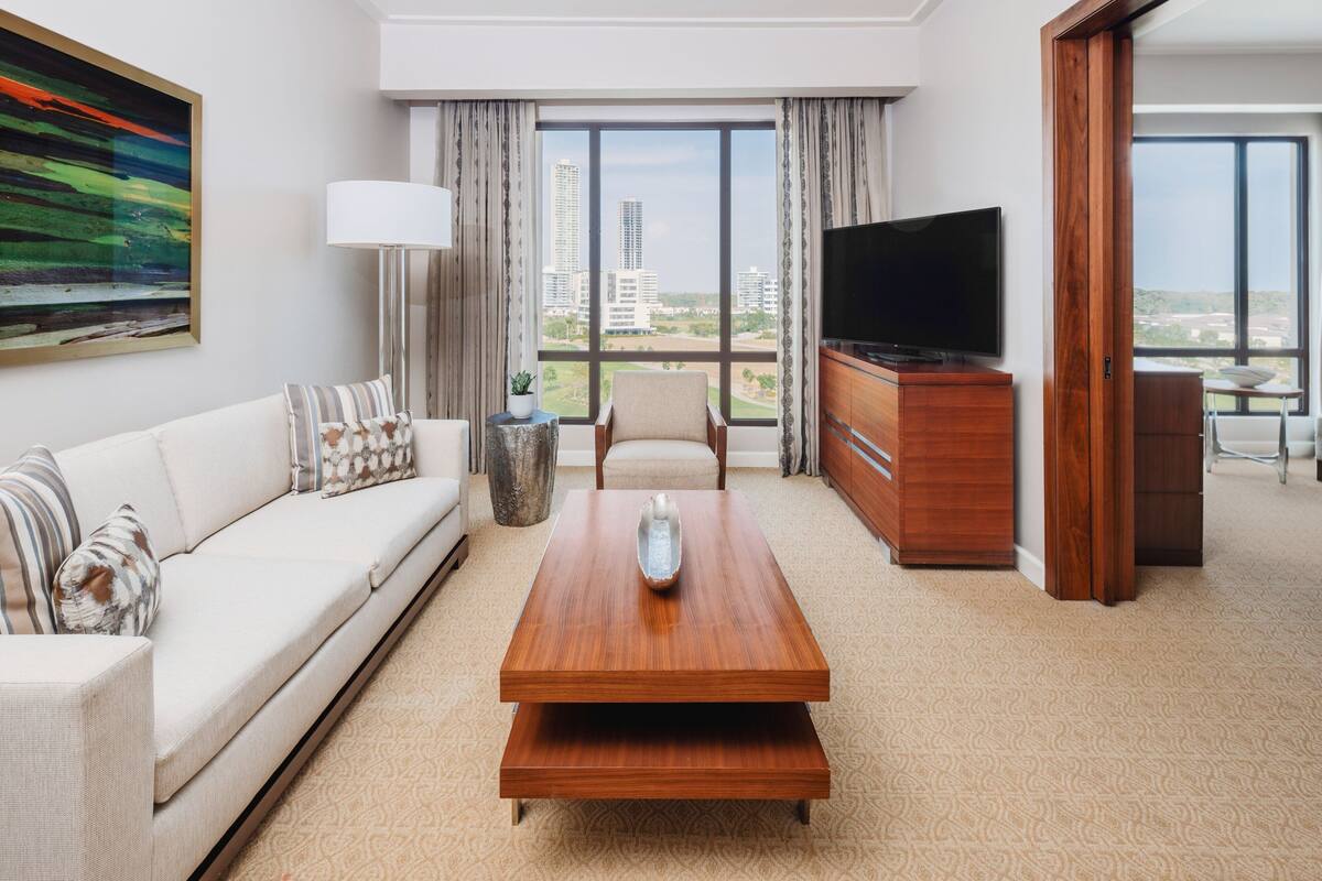 A modern hotel room with a sofa, a wooden coffee table, a TV, and a view of the cityscape.