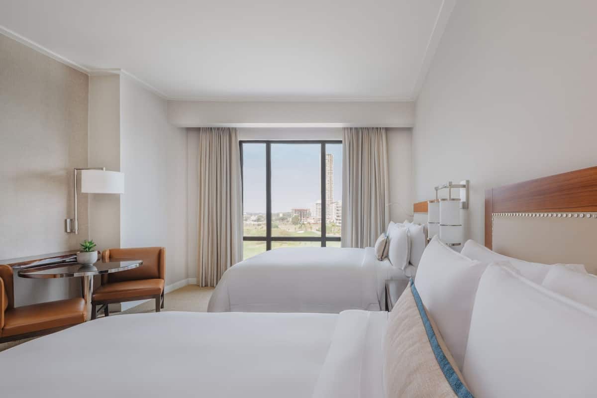 A hotel room with two beds, a desk, a chair, a lamp, and a view of the city.