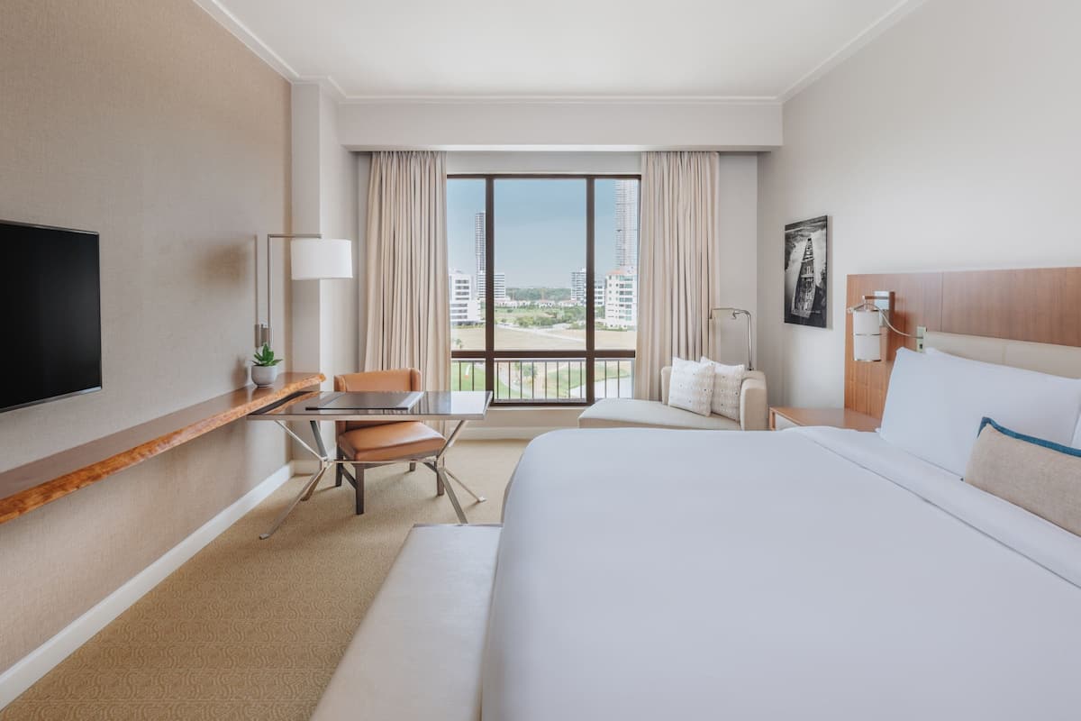 A hotel room with a large bed, a desk with a chair, a TV, and a window with a city view.