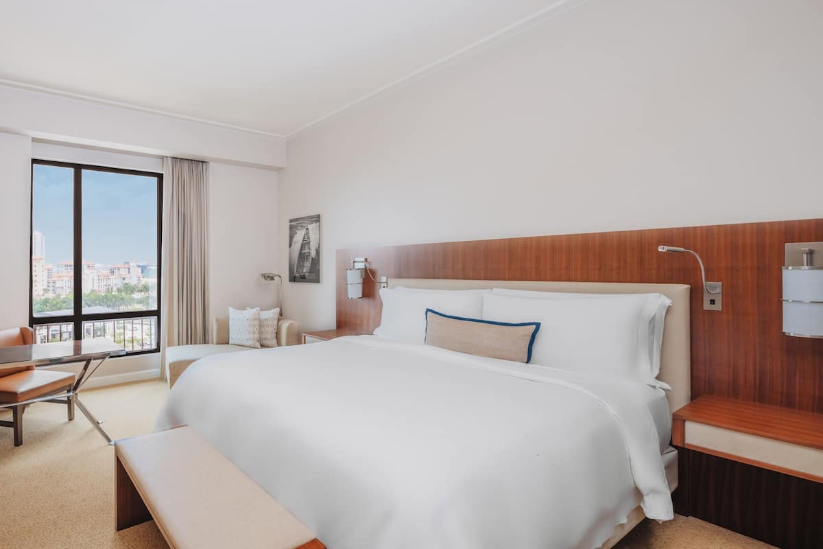 A hotel room with a large bed, a desk, and a chair. There is a window with a view of buildings.