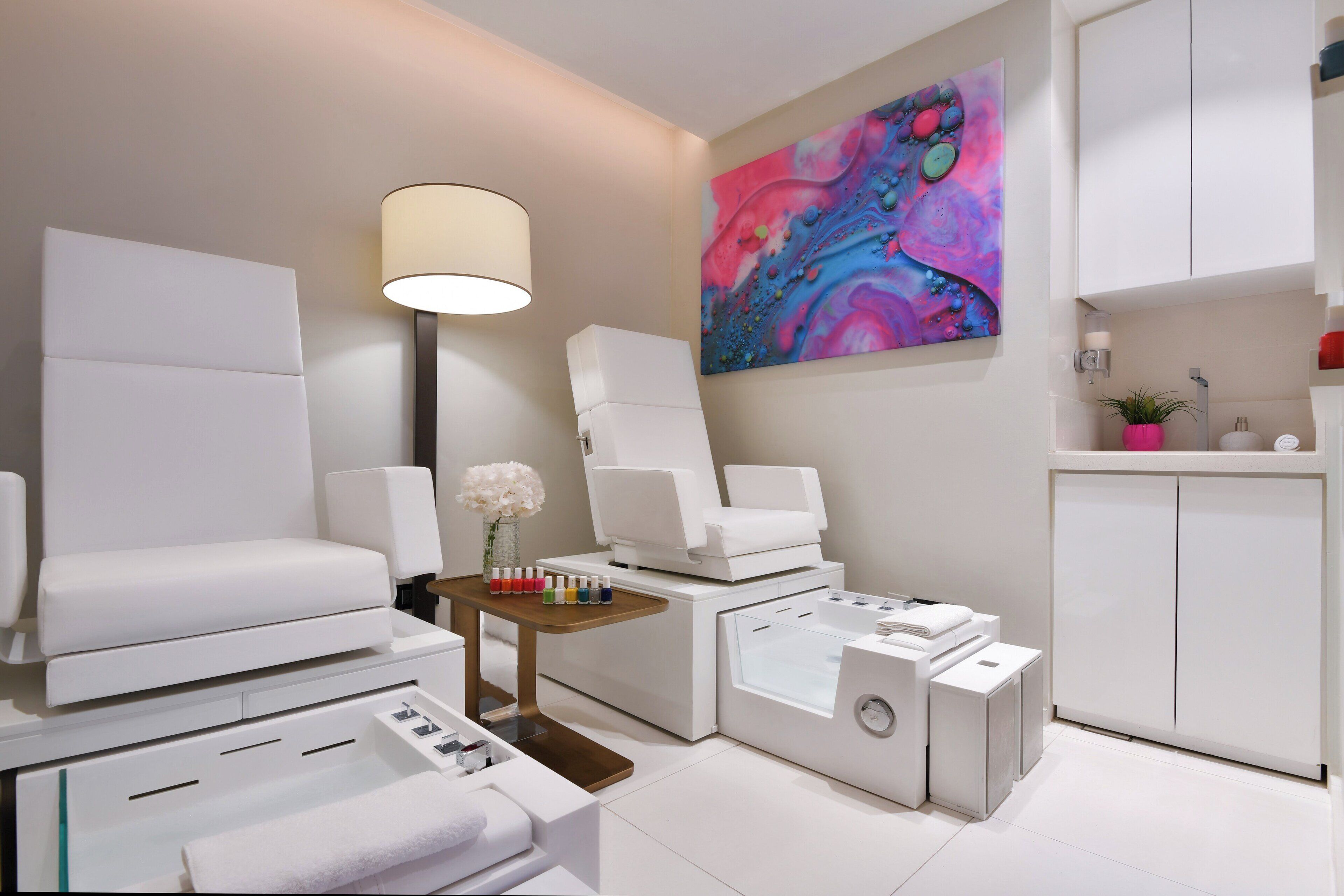 A modern spa room with a large white sofa, a massage table, a foot bath, and a colorful abstract painting on the wall.