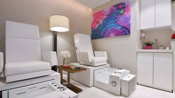 A modern spa room with a large white sofa, a massage table, a foot bath, and a colorful abstract painting on the wall.