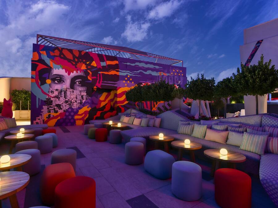 An outdoor seating area with round tables and colorful cushions, a vibrant mural on the wall, and a clear sky.
