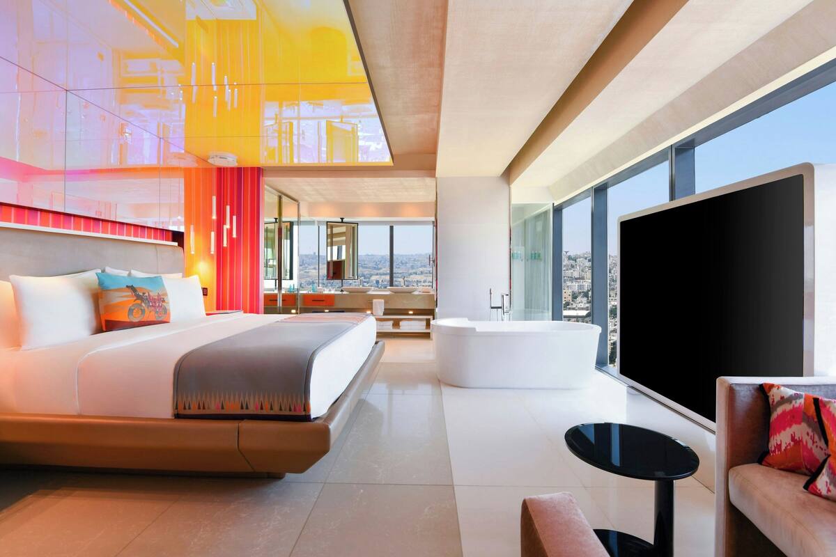 A modern hotel room with a large bed, a sitting area with a sofa and a small table, a bathtub, and a large window offering a city view.