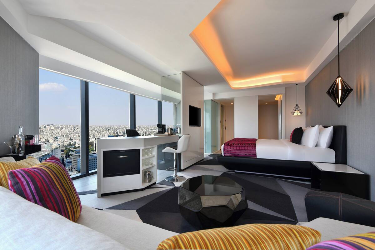 A modern hotel room with a large window offering a city view, a bed, a desk with a chair, and a glass coffee table.