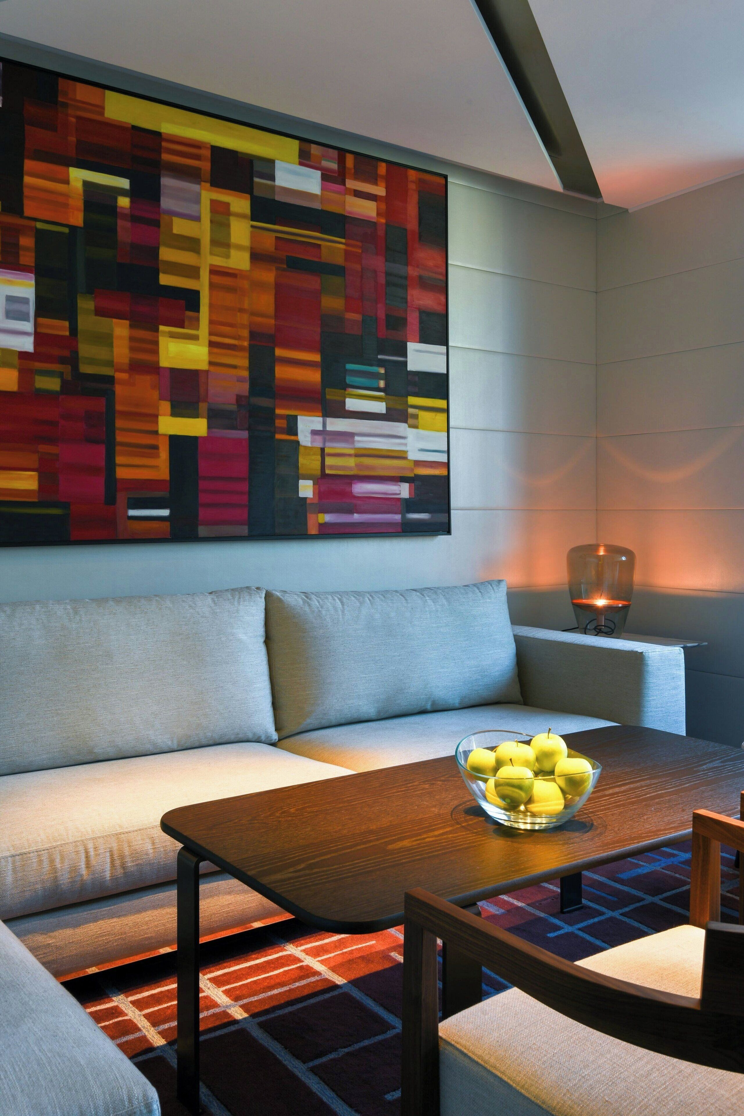 Un salon moderne avec un canapé, une table basse en bois sur laquelle se trouve un saladier rempli de fruits, et un tableau abstrait aux couleurs vives accroché au mur.
