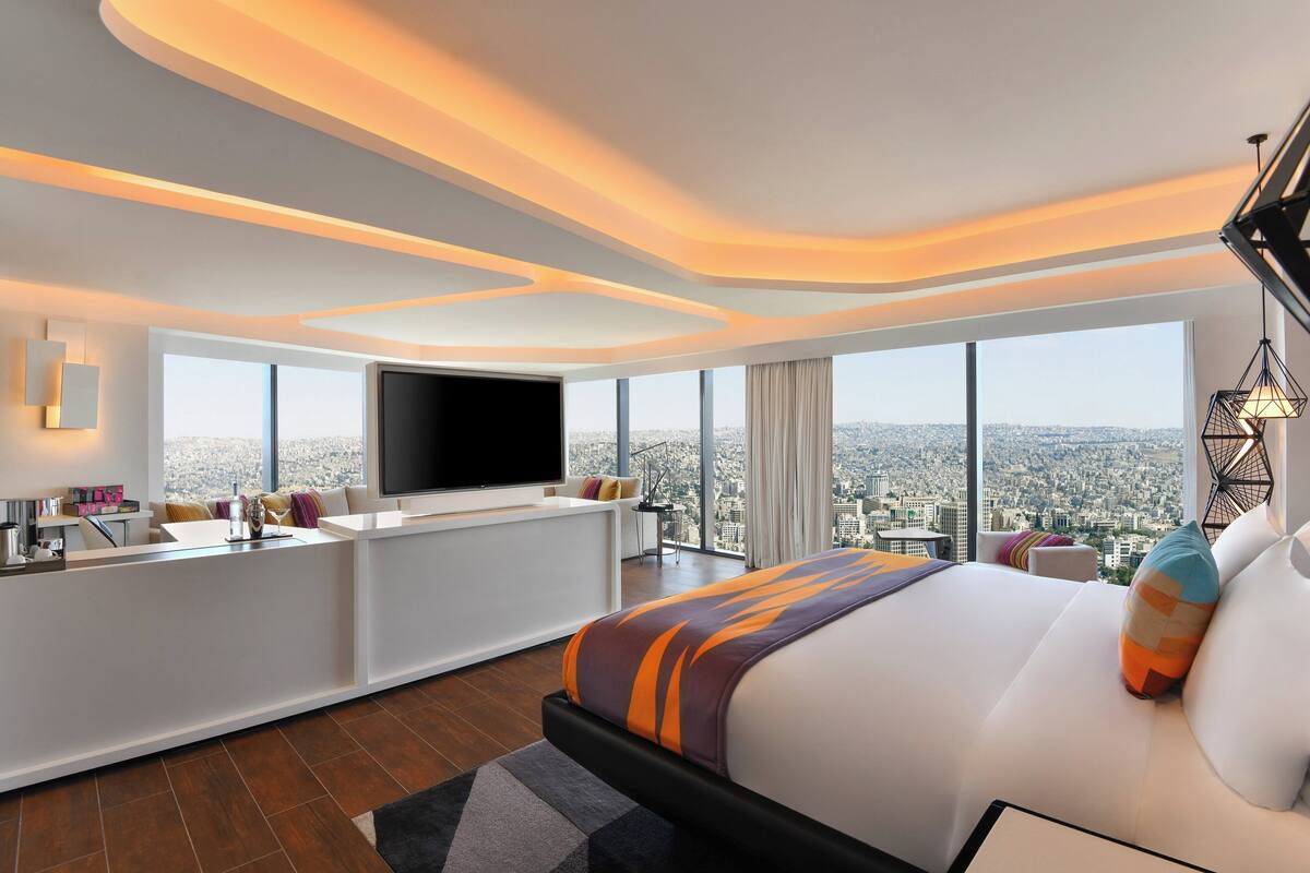 A modern hotel room with a large bed, a TV, a desk, and a view of the city.