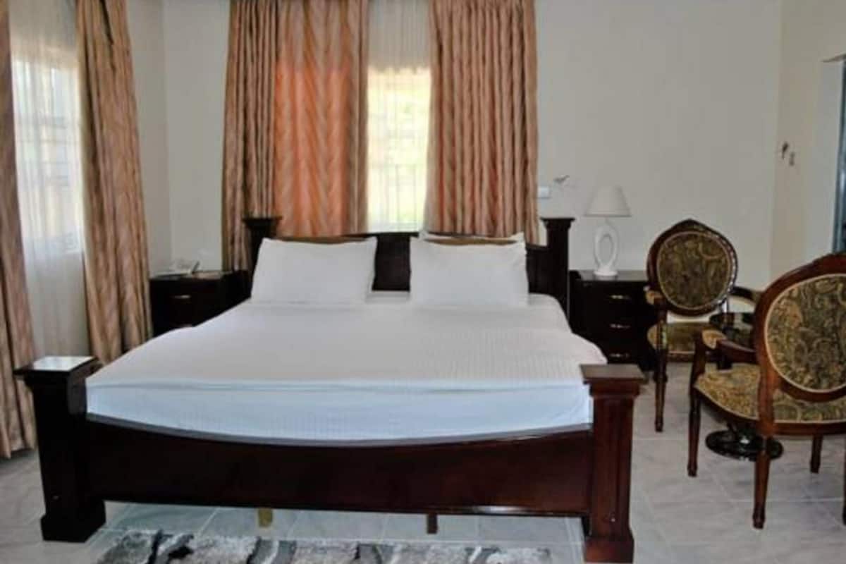 A large bed with a wooden frame, a bedside table, a chair with a patterned cushion, and curtains on the windows.