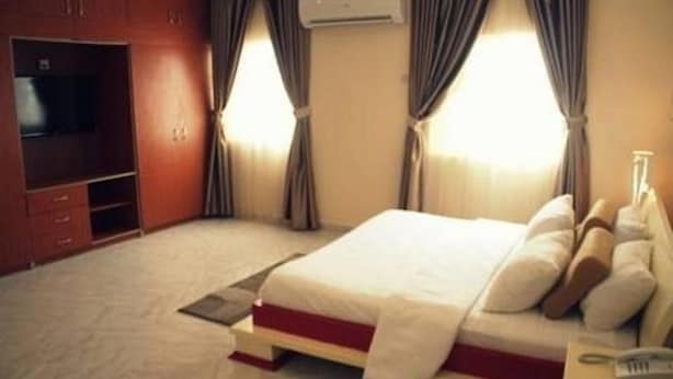 A hotel room with a bed, a TV, a wardrobe, and an air conditioning unit. - Chateau De Trybze (Abuja)