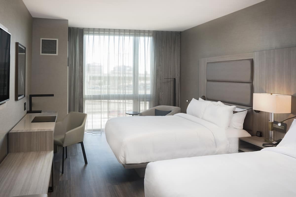 Habitación de hotel con dos camas, un escritorio, una silla, un televisor y un ventanal grande con cortinas.