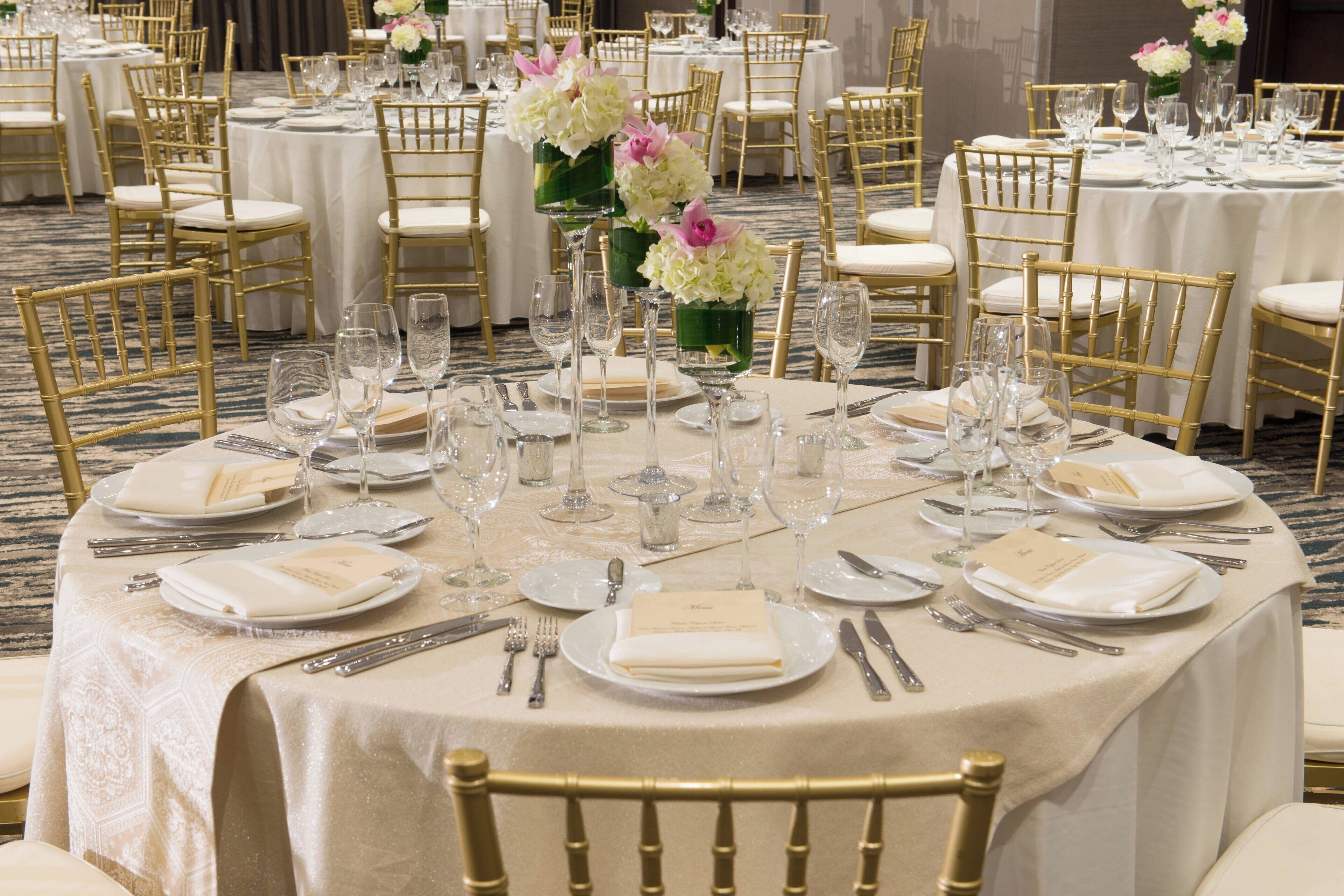 Ein Bankettsaal mit runden Tischen, gedeckt für ein Essen, jeweils mit weißen Tischdecken, goldenen Stühlen und floralen Mittelstücken.