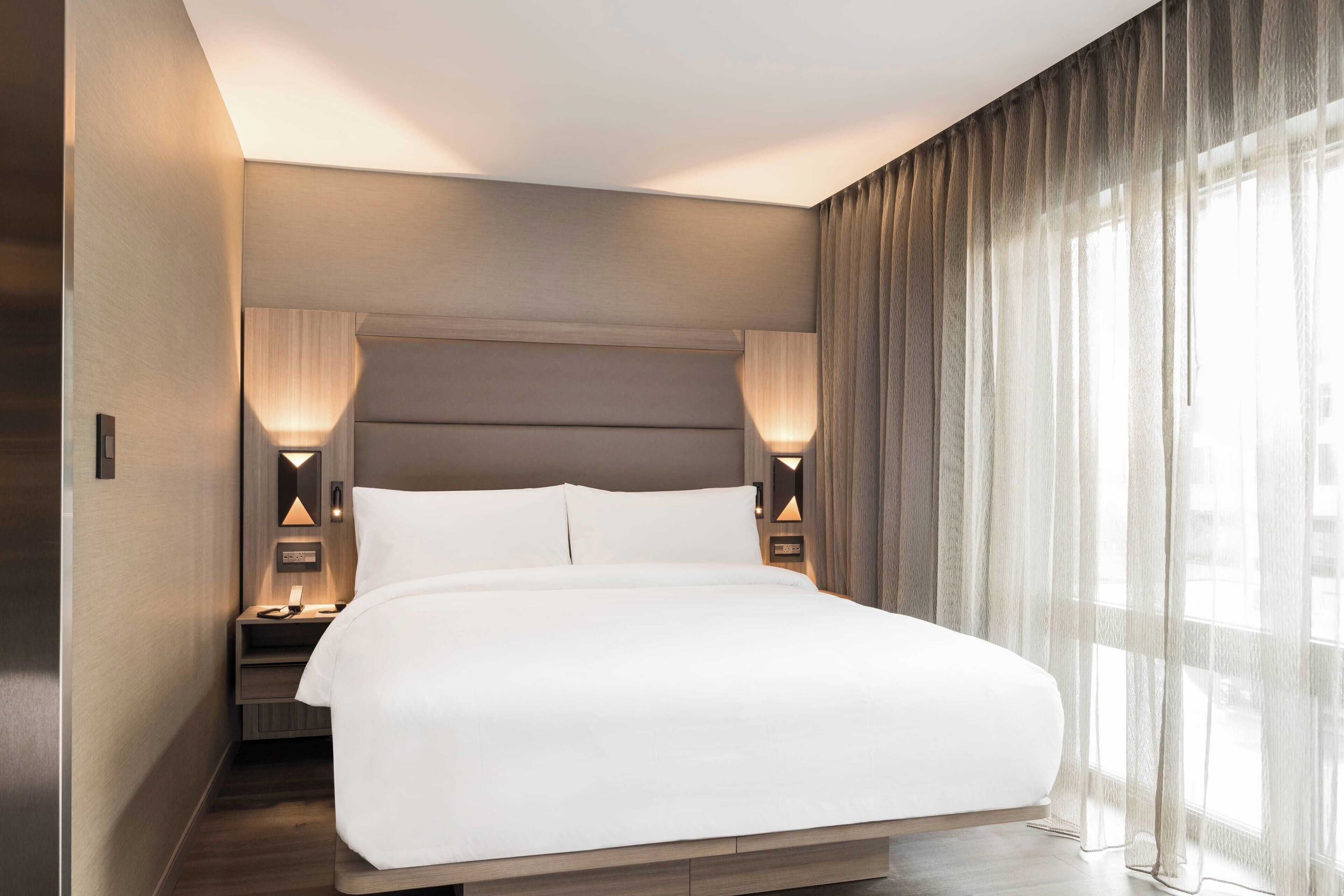 Una habitación de hotel moderna con una cama grande, dos mesitas de noche con lámparas y vistas a través de cortinas translúcidas.