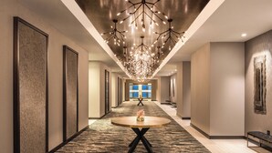 A modern hotel corridor with a central round table, a unique chandelier, and a view to a large window at the end.