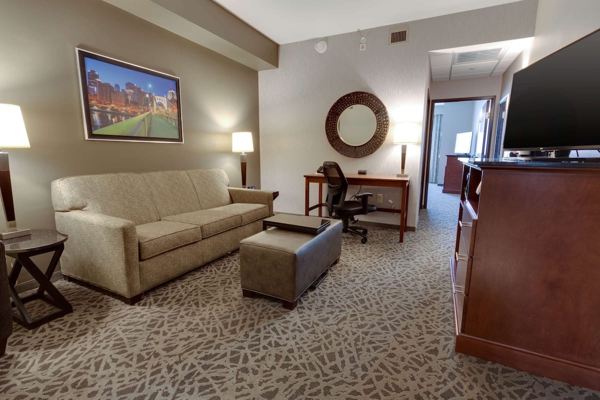 A hotel room with a sofa, ottoman, desk, chair, and a television.
