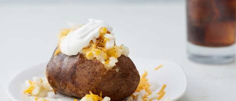 A baked potato topped with sour cream and shredded cheese, served on a white plate with a glass of iced tea in the background.