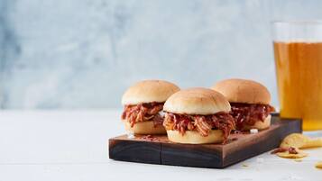 Due panini con pulled pork su un tagliere di legno, con un bicchiere di birra sullo sfondo.