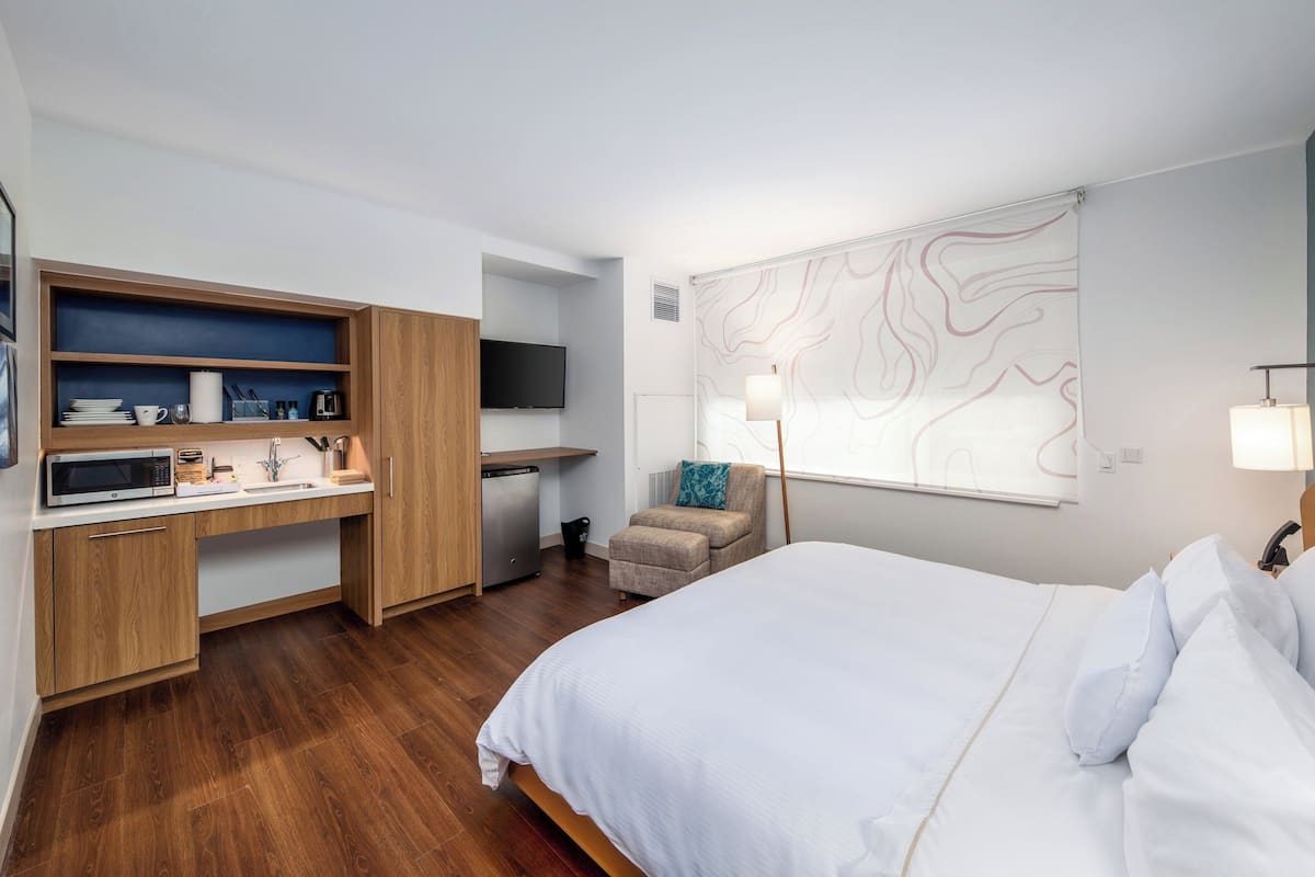 A compact hotel room with a bed, a desk with a microwave, a TV, and a wall-mounted lamp.