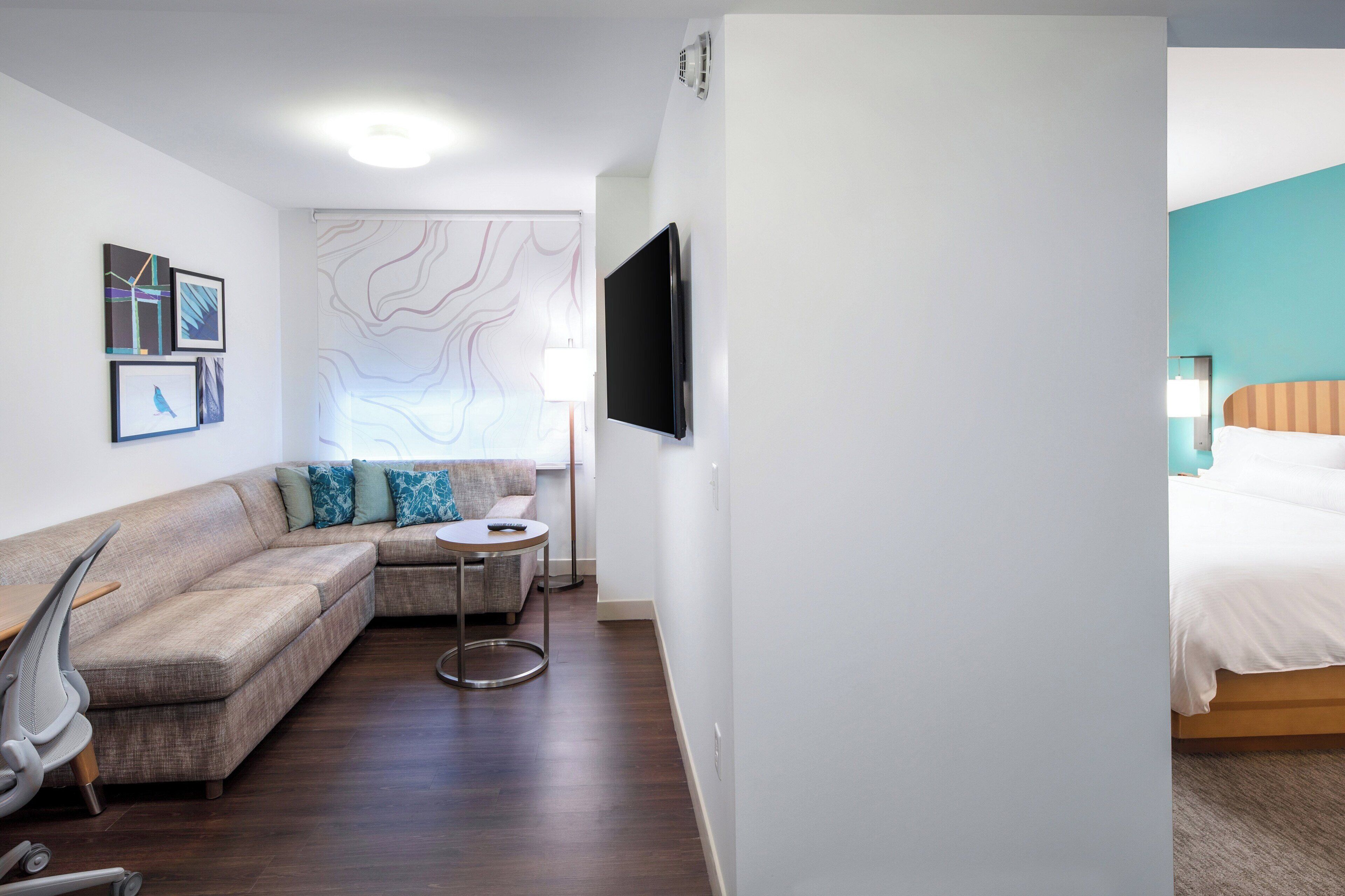 A modern hotel room with a sofa, a small round table, a TV mounted on the wall, and a bed with a headboard.