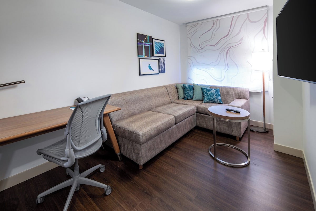 A modern living room with a grey office chair, a grey sofa, a small round table, and a large window with a view.