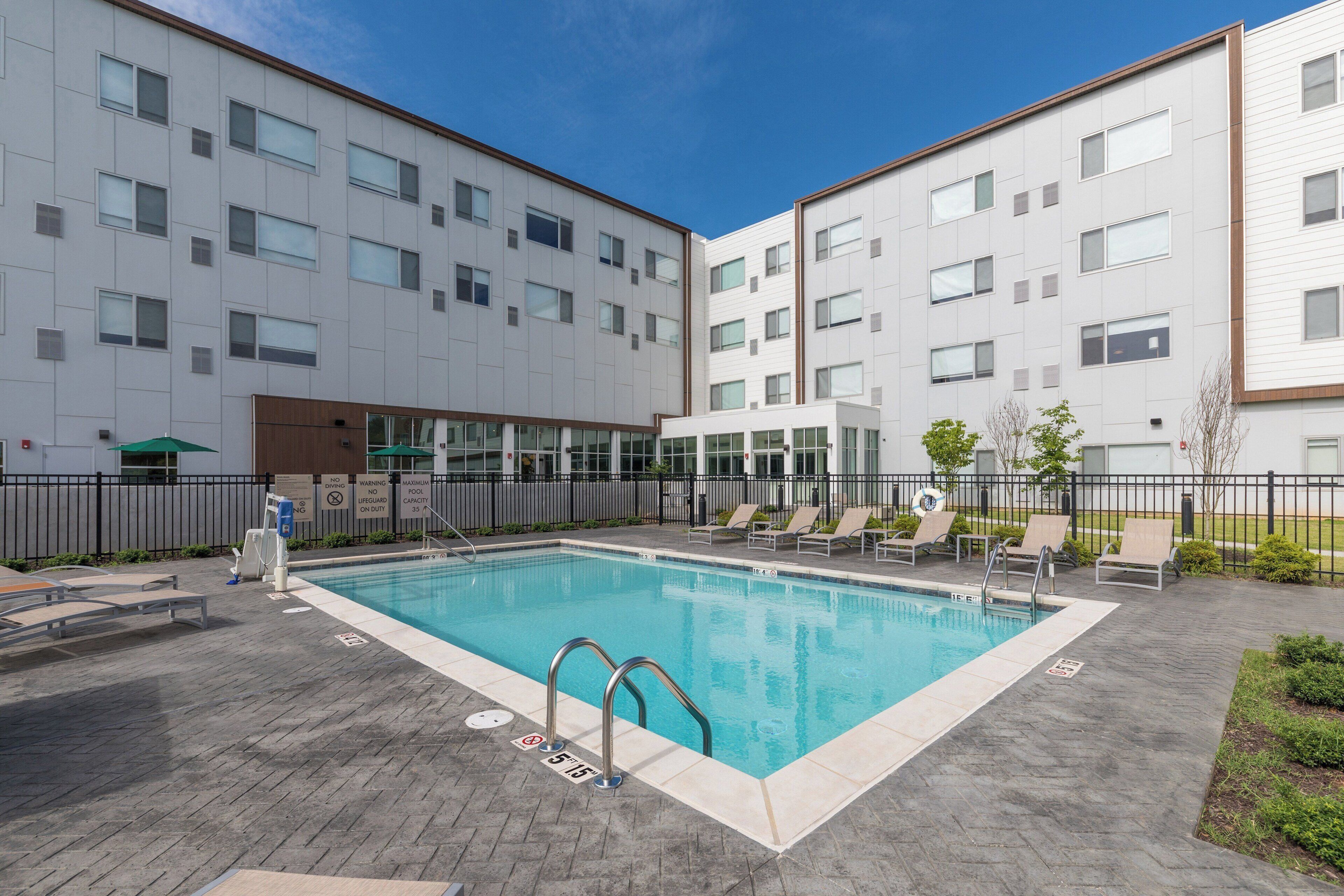 A swimming pool with a wooden deck, lounge chairs, and a building with multiple windows.