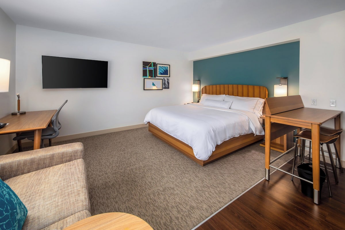 A hotel room with a bed, a desk, a chair, a TV, and artwork on the wall.